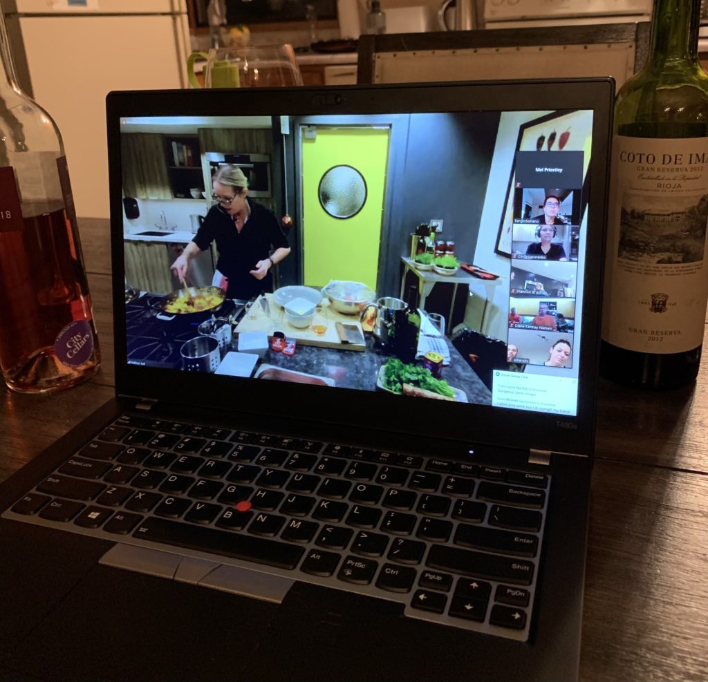 a laptop showing a cooking demonstration sitting on a table beside some wine bottles